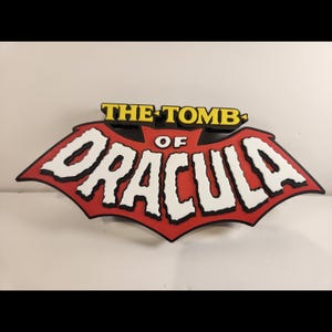 May include: A three-dimensional wall sign featuring the text THE TOMB OF DRACULA. The word DRACULA is in large, jagged white letters on a red bat-shaped background, while THE TOMB is in yellow letters above it with black outlines.