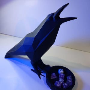May include: A black, geometric, low-poly raven figurine with an open beak. The bird stands on a circular, black, textured nest-like base that holds several purple translucent polyhedral dice and one white six-sided die with black pips.