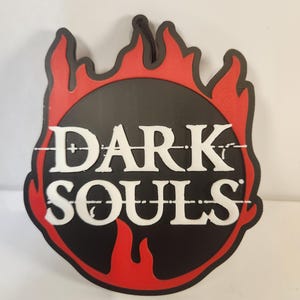 May include: A black and red emblem featuring the text "DARK SOULS" in white. The emblem is in the shape of a flame, with a black circle at the centre. The text is positioned within the circle.