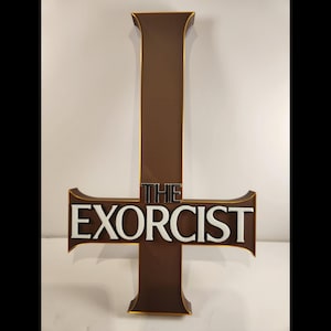 Könnte beinhalten: Ein braunes, kreuzförmiges Wandschild mit dem Text THE EXORCIST in fetten, weißen, erhabenen Buchstaben. Das Kreuz hat einen dünnen goldfarbenen Rand an den Kanten, und das Wort THE steht in kleineren schwarzen Buchstaben über dem Haupttitel.