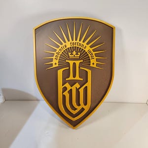 May include: A shield-shaped emblem with a brown background and gold-colored trim and details. The emblem features the text "audentes fortuna iuvat" in a sunburst design above a stylized letter "R" and "W".