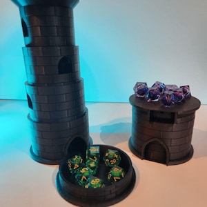 May include: Two black 3D printed dice towers, one tall and one short, with a removable lid. The tall tower has three levels and the short tower has one level. Both towers have a small opening on the side for dice to be rolled out. The tall tower has green dice inside and the short tower has purple dice inside.