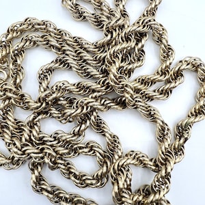 May include: A gold-toned rope chain necklace with a thick, twisted design. The chain is made of multiple strands of metal that are woven together to create a rope-like appearance.