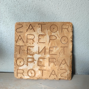 May include: A square, tan-colored tile with the Latin phrase "SATOR AREPO TENET OPERA ROTAS" carved into it. The letters are arranged in a square pattern, with the same letters appearing in each row and column.