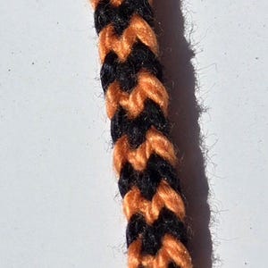 May include: A close-up of a black and orange braided cord. The cord is made of two strands of yarn, one black and one orange, that are woven together in a simple pattern.