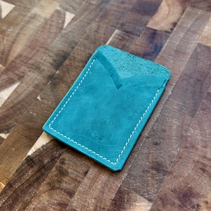 May include: Teal leather card holder with white stitching. The card holder has a V-shaped opening at the top and is rectangular in shape. The leather has a suede-like texture.