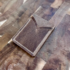 Könnte beinhalten: Ein brauner Lederkartenhalter mit strukturierter Oberfläche und weißen Nähten. Der Kartenhalter hat ein schlichtes, minimalistisches Design mit einer V-förmigen Öffnung. Der Artikel ist wahrscheinlich handgefertigt und für Kreditkarten oder Visitenkarten konzipiert.