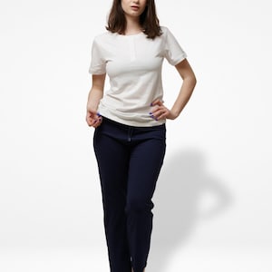 May include: A woman in a white short-sleeved top with a buttoned neckline and navy blue trousers with a drawstring. The casual outfit is suitable for everyday wear. The woman is standing against a white backdrop.