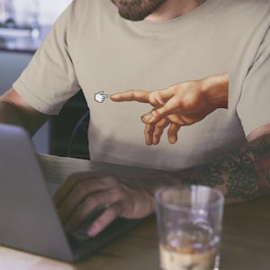 Könnte beinhalten: Beiges T-Shirt mit einem pixeligen Cursor, der auf eine Hand zeigt, ein Detail aus Michelangelos Die Erschaffung Adams. Die Person sitzt an einem Tisch mit einem Laptop und einem Glas Eiskaffee.
