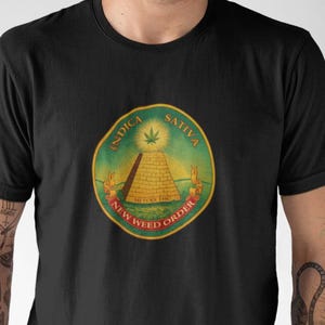Könnte beinhalten: Schwarzes T-Shirt mit einem kreisförmigen Design. Das Design zeigt die Wörter "Indica Sativa" und "New Weed Order" um eine Pyramide und ein Cannabisblatt. Die Grafik verwendet grüne, gelbe und rote Farben.