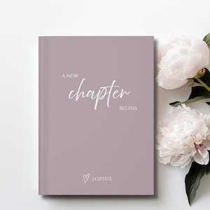 May include: A mauve hardback book with the white text A NEW CHAPTER BEGINS and a small heart icon above the name SOPHIE printed on the front cover. Two large, pale pink peony flowers with green leaves are positioned to the right of the book.