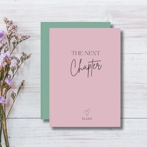 May include: A pink journal with the words "THE NEXT Chapter" in black script. Below the text is a small heart and the name "ELLEN". A green journal is behind the pink journal. The background is a white wooden surface with purple flowers.