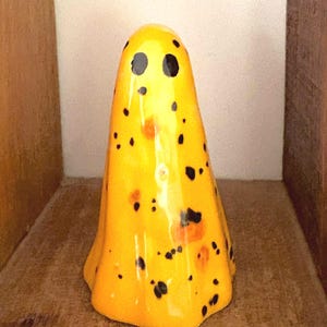 Op de afbeelding: Een gele, spookvormige keramische figuur met zwarte en oranje spikkels. Het beeldje heeft twee zwarte stipogen en een afgeronde bodem. De geest wordt tentoongesteld op een houten plank.