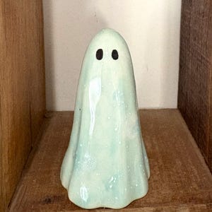 May include: A ceramic ghost figurine with a light blue glaze and black eyes. The ghost is standing on a wooden shelf.