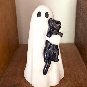 Peut inclure: Figurine de fantôme en céramique blanche avec des yeux noirs, tenant un chat noir. Le fantôme a une forme simple et arrondie, et le chat est brillant et détaillé. Décoration sur le thème d'Halloween pour une étagère ou une cheminée.