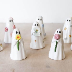 May include: A collection of white ceramic ghost figurines, each with black dot eyes and holding various items like flowers and gifts. Some ghosts have painted floral details. The figurines are arranged on a light-colored surface.