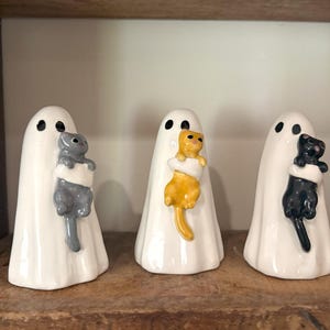 Peut inclure: Trois figurines de fantômes en céramique blanche, chacune tenant un chat de couleur différente. Les chats sont gris, jaunes et noirs. Les fantômes ont des yeux en points noirs et sont exposés sur une étagère en bois.