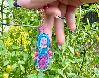 May include: A blue and pink flip phone charm with a cartoon bunny on the screen. The charm has a pink flower and glitter accents.