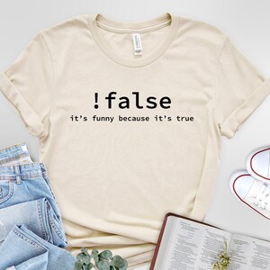 May include: A beige t-shirt with black text that reads "!false it's funny because it's true". The shirt is lying on a white surface with a pair of blue jeans and a book.