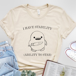 May include: A beige t-shirt with a black and white illustration of a penguin holding a knife. The text "I have stability (ability to stab)" is printed on the shirt.