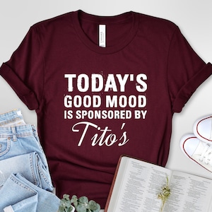 May include: A maroon t-shirt with white text that reads "Today's Good Mood Is Sponsored By Tito's".