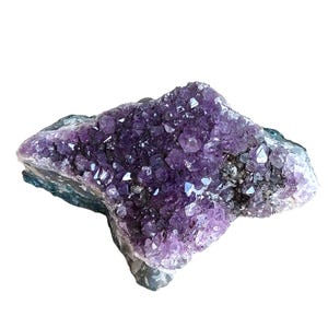 May include: A large, raw amethyst crystal cluster. The crystal features a deep purple hue with sparkling, pointed crystal formations. The base of the cluster has a dark, greyish-green color. The overall shape is irregular and natural.
