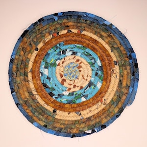 Pode incluir: Tapete redondo feito à mão com um design em espiral. Apresenta anéis concêntricos de tecido em tons de azul, castanho e bege. O centro tem um padrão floral azul claro e bege, e a borda externa é azul escuro. O diâmetro é de cerca de 30 cm.