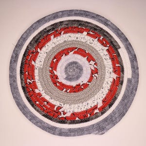 May include: Circular braided rug with a spiral design. The rug features concentric rings of red, white, gray, and black fabric. The outer edge is a solid gray, and the rug is made of stitched fabric strips.