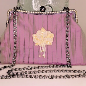 Puede incluir: Un bolso de mano de tela plisada de color morado con una correa de cadena plateada y un adorno de flor de cuentas blancas.