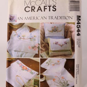 Puede incluir: Un sobre de patrón de costura McCall's Crafts con diseños bordados. El sobre muestra imágenes de fundas de almohadas bordadas, un mantel y otros artículos con motivos florales y de pájaros. El texto "AN AMERICAN TRADITION" es visible.