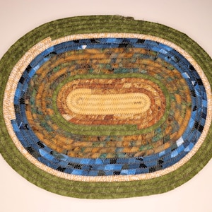Pode incluir: Tapete oval trançado com borda verde, detalhe creme e um design central de tecidos azuis, castanhos e amarelos. O tapete é feito à mão e apresenta uma construção costurada. O design é uma mistura de cores e texturas.