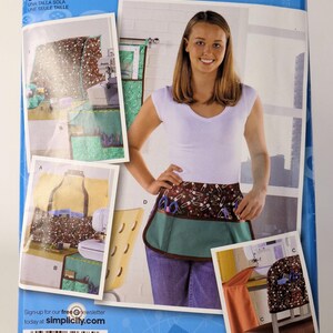 May include: Simplicity 2679 Project Runway sewing pattern envelope featuring a multi-pocket apron with a brown safety pin print and solid teal fabric. The packaging displays various craft storage organizers and chair covers with matching patterns.