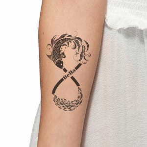 Peut inclure: Un tatouage noir représentant un poisson koï avec des détails fluides, intégré dans un symbole infini. Le mot "Bella" est écrit sur le symbole. Un banc de petits poissons complète le motif.