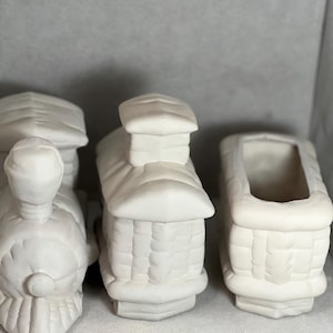 Puede incluir: Tres vagones de tren de cerámica blanca, listos para ser pintados. El primer vagón es una locomotora con un frente redondeado y una chimenea. El segundo vagón tiene un frente cuadrado y una chimenea. El tercer vagón es un vagón de carga con una abertura rectangular.