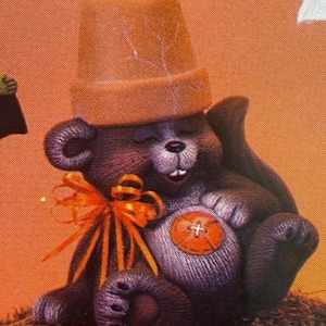 May include: A dark brown bear figurine with a light brown terracotta flower pot on its head. The bear has a bright orange ribbon tied around its neck and a large orange button sewn onto its belly. The bear is sitting with its eyes closed.