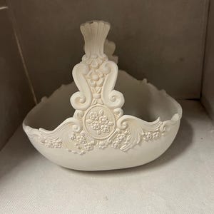 May include: Off-white decorative basket with ornate floral and scroll detailing. The basket features a handle and a rounded bowl shape, with embossed floral patterns around the rim and on the handle. The overall design is classical and elegant.