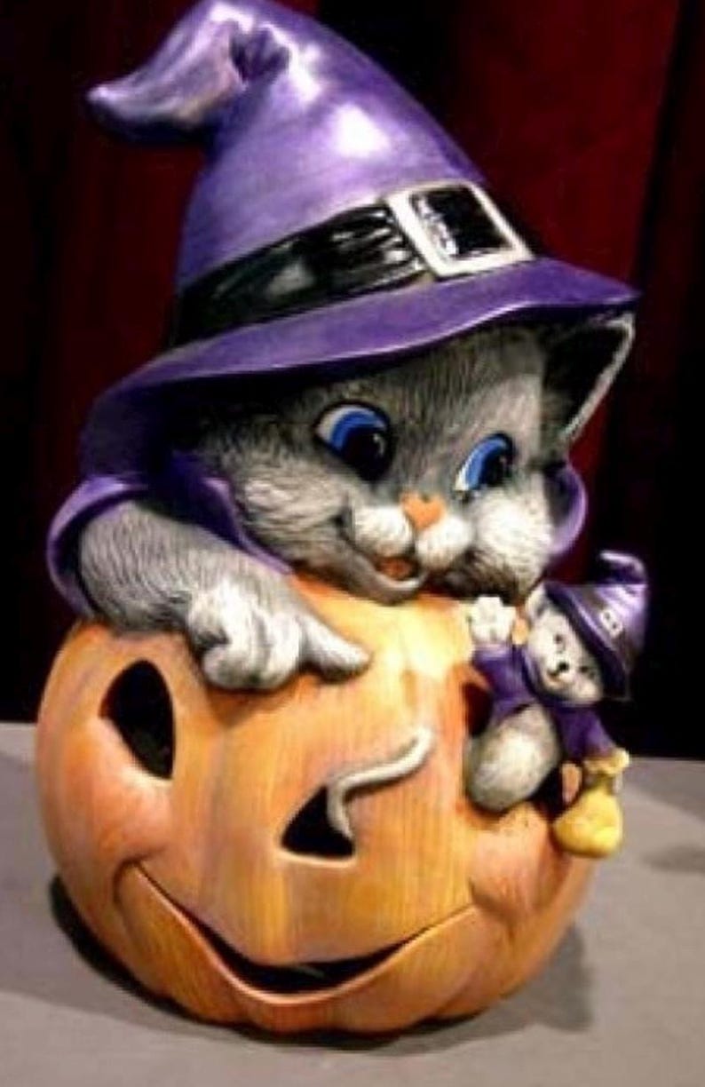 May include: A Halloween-themed ceramic figurine featuring a gray cat wearing a purple witch hat, perched atop an orange pumpkin with a carved face. A smaller cat figurine is also present. The figurine has blue eyes and is painted with detail.