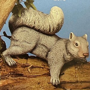 Könnte beinhalten: Eine graue Eichhörnchenfigur mit strukturiertem, buschigem Schwanz und detailliertem Fell, positioniert auf einem braunen, strukturierten Baumstamm. Das Eichhörnchen hat eine helle Nase, wache Augen und kleine Ohren, dargestellt in einer realistischen Pose.