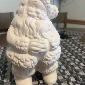 May include: A white ceramic Santa Claus figure. The figure has a smiling face, a long beard, and fluffy hat and coat trim. The Santa Claus figurine is a festive decoration.