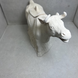 Puede incluir: Figura de vaca de cerámica blanca con una silla de montar en la espalda. La vaca está de pie sobre sus cuatro patas y tiene una cara detallada.