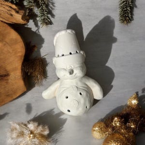 May include: White ceramic snowman figurine with a hat and a smiling face. The snowman has a round body with three buttons and a carrot nose.