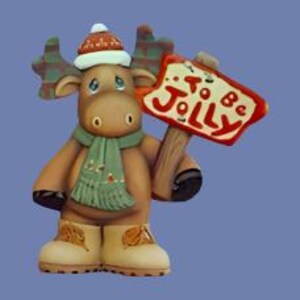 May include: A brown moose figurine wearing a red and white winter hat, a green scarf, and tan boots. The moose holds a wooden sign with red text that reads To Be Jolly. The figurine features large antlers and a smooth, matte finish throughout.