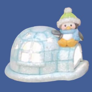 Op de afbeelding: Een grillig beeldje van een pinguïn met een groene muts en blauwe sjaal, bovenop een lichtblauwe iglo. De iglo heeft een raam en is bedekt met glitter, wat een winters tafereel creëert. De pinguïn heeft gele voeten en een witte buik.