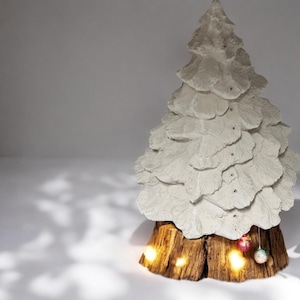 May include: A white concrete Christmas tree with a rustic wood base and small lights. The tree is decorated with red and green ornaments.