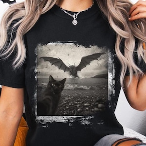 May include: Black t-shirt with a graphic print of a black cat looking at a large, winged creature with tentacles emerging from the water. The graphic is in a distressed, vintage style.