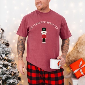 Puede incluir: Una camiseta granate con la inscripción "NUTCRACKER SURVIVOR" sobre un soldado cascanueces de dibujos animados. La persona lleva un pantalón de pijama a cuadros rojos y negros y sostiene una taza blanca con un diseño de Papá Noel. Un árbol de Navidad y un regalo envuelto están en el fondo.