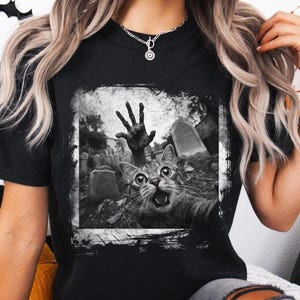 May include: Black and white graphic t-shirt featuring a cat with a zombie hand reaching out of a grave. The cat has a surprised expression and its mouth is open.