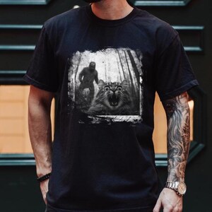 May include: Black t-shirt with a graphic print of a cat looking scared with a blurry figure of a monster behind it.