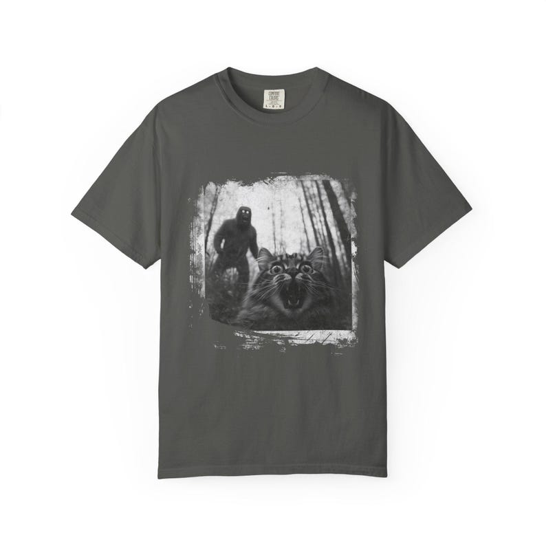 May include: A dark gray t-shirt with a black and white image of a cat looking scared with a figure of a large hairy creature behind it in the woods.
