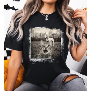 May include: Black t-shirt with a distressed white frame around a black and white photo of a cat looking surprised with a unicorn in the background.